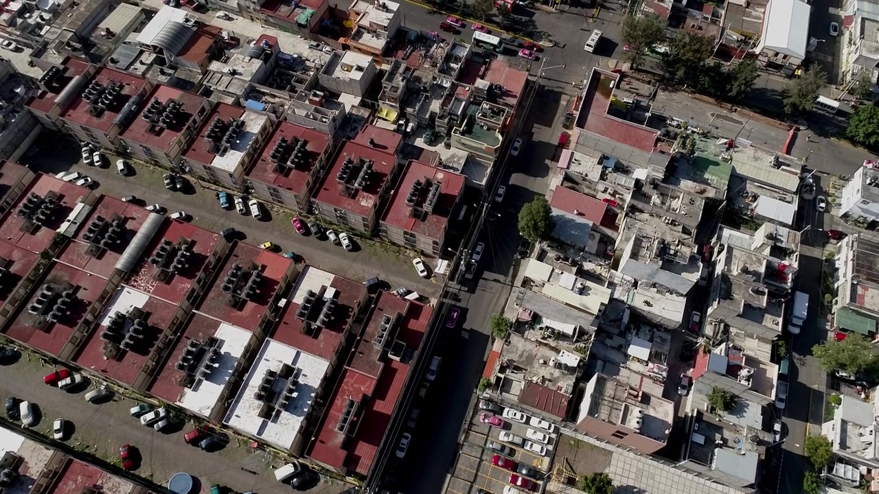 Ripresa aerea con drone di un denso quartiere residenziale con edifici compatti, strade strette, veicoli parcheggiati e sviluppo urbano in una giornata di sole.