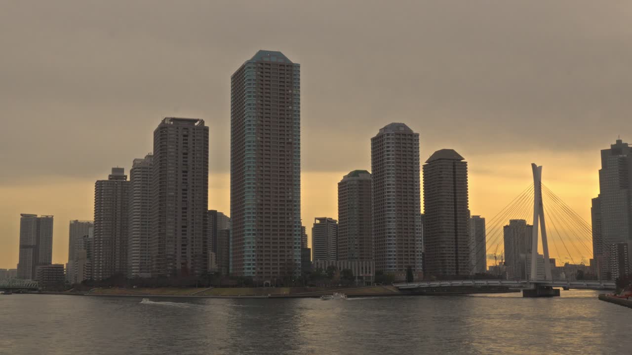 Uma vista panorâmica do Rio Sumida em Tóquio ao pôr do sol, apresentando o horizonte de River City 21 e uma ponte ao longe.