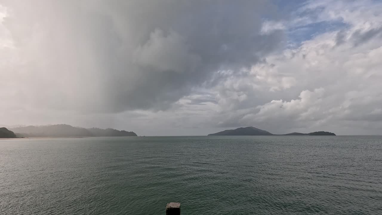 A wide shot captures a dramatic rainstorm moving across the sea toward a forested coastline on Ko Phayam, with shifting clouds and changing light
