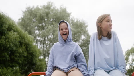 Little girl with down syndrome sitting on the top of a children&#039;s park with her blonde friend on a windy day