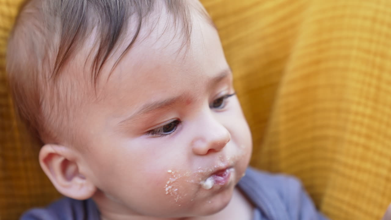 Baby boy eating porridge from spoon. Mother giving food to the child sitting in a yellow chair. Close up.