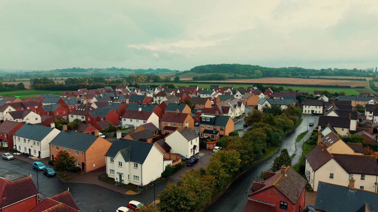 Boligområde med rækker af huse omgivet af grønne marker og træer i udkanten af byen under en skyet himmel, fredeligt forstadssamfund med vejnetværk synligt ovenfra