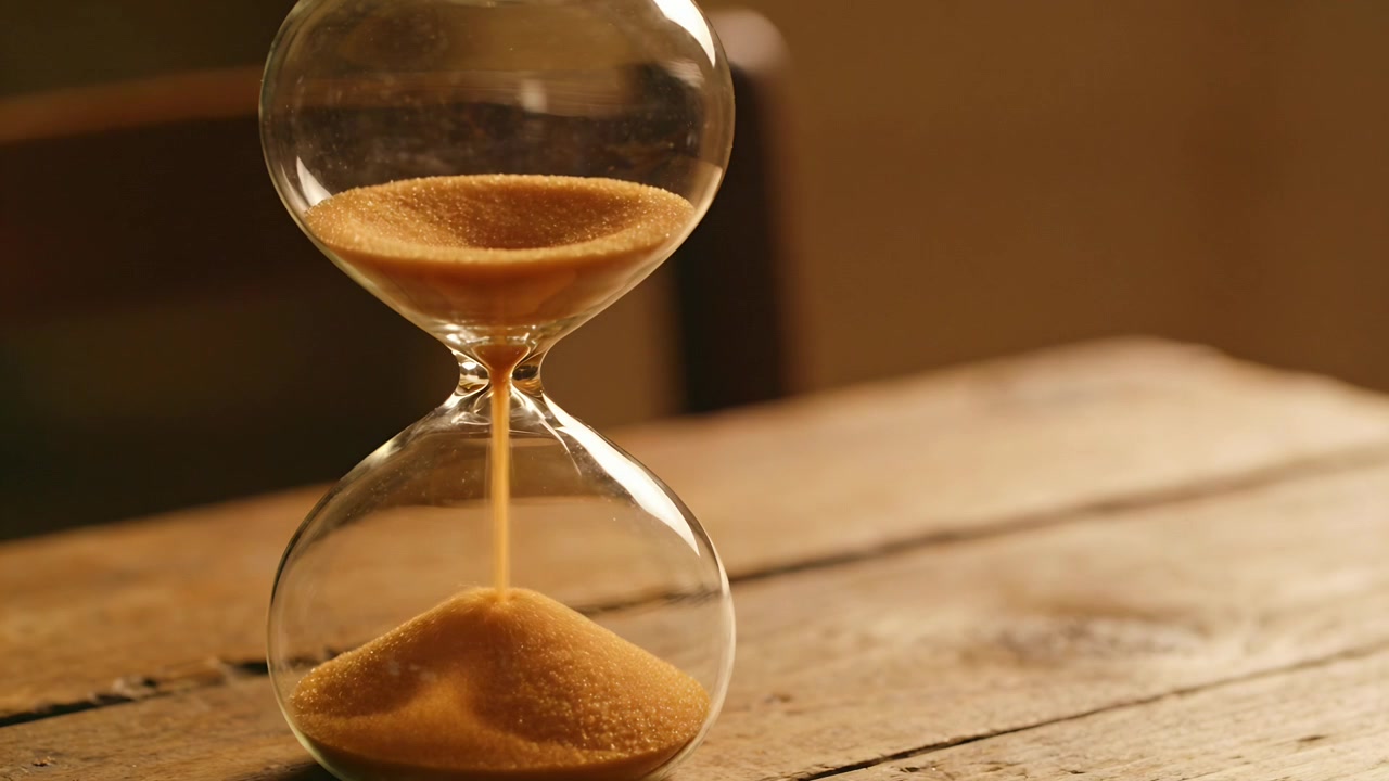Recording glass hourglass pouring amber sand on wooden tabletop, indicating time passing