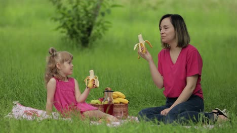 Family-weekend-at-picnic.-Daughter-child-girl-with-mother-on-grass-meadow-eating-bananas,-having-fun