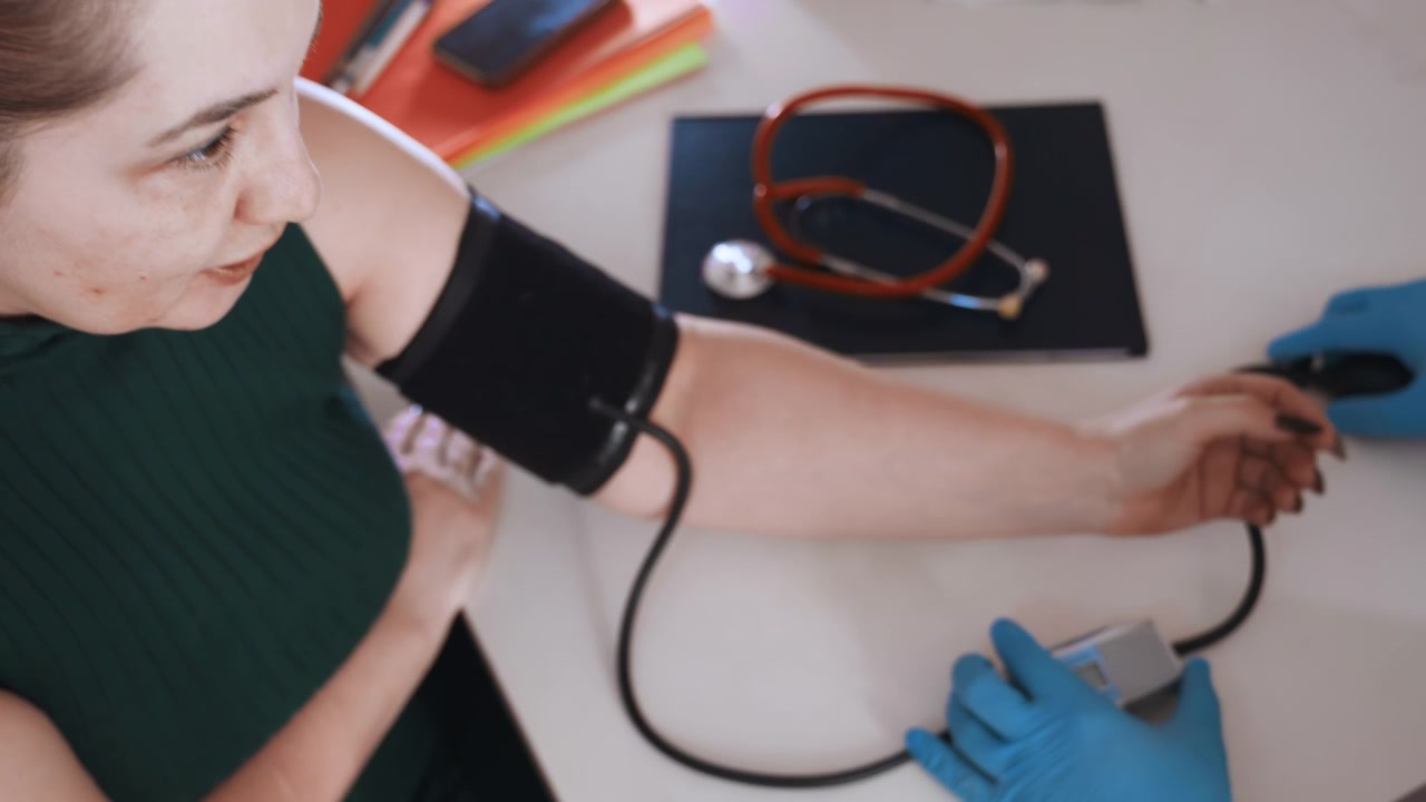 Applicazione ravvicinata del bracciale indossando guanti, mostrando braccio e dispositivo, posizionamento attento del bracciale nero, tubi e manometro visibili sulla scrivania, tecnica clinica e igiene concentrate, mani guantate che regolano la vestibilità