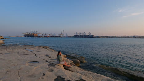 a lady sits on the rocks on the mediterranean coast, with the port of valletta in the background