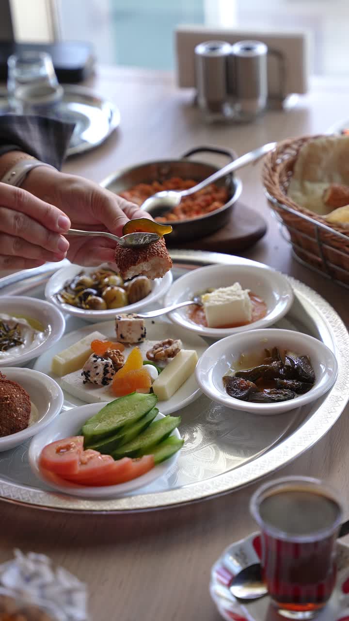 Mani che aggiungono miele a pane al sesamo sopra un piatto tradizionale per la colazione con formaggio, olive, verdure e tè in un accogliente ristorante al coperto durante il giorno