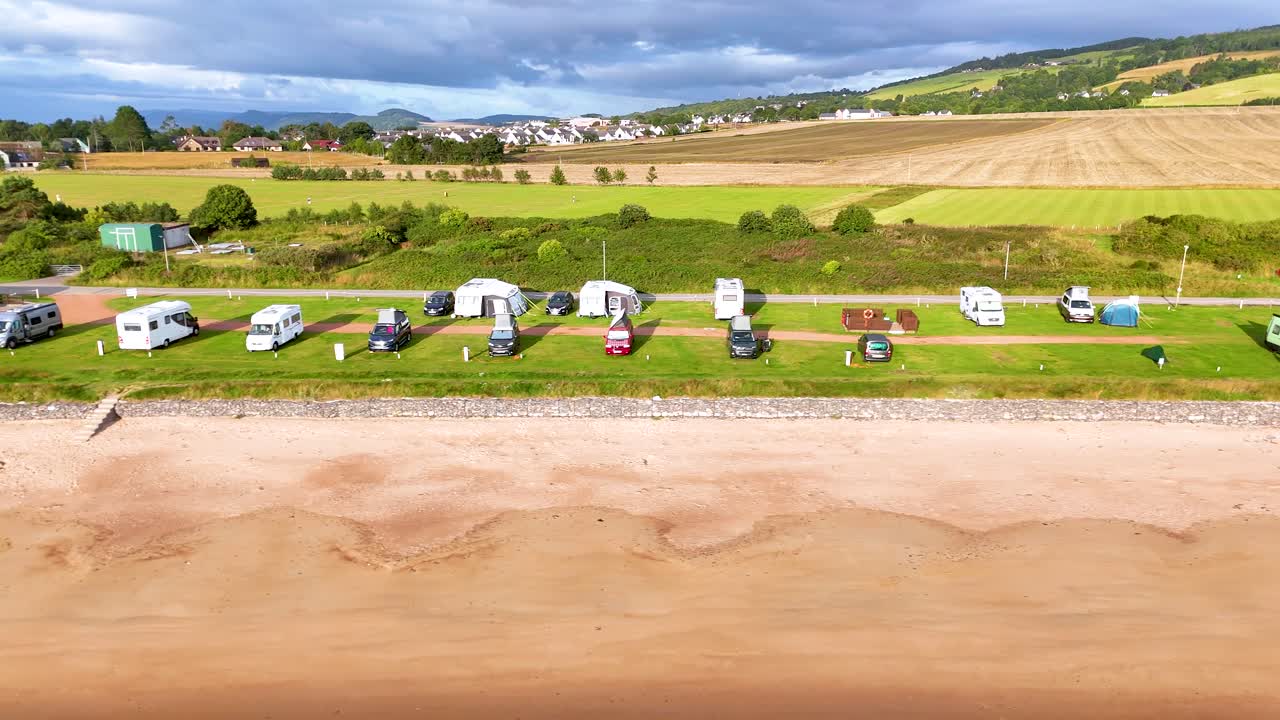 Aerial footage smoothly pans across a coastal caravan park in Rosemarkie, Scotland, revealing campers, grassy fields, and sandy shoreline under bright daylight
