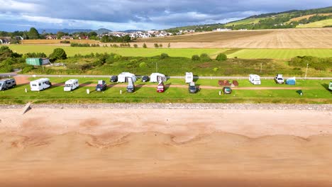 aerial footage smoothly pans across a coastal caravan park in rosemarkie, scotland, revealing campers, grassy fields, and sandy shoreline under bright daylight