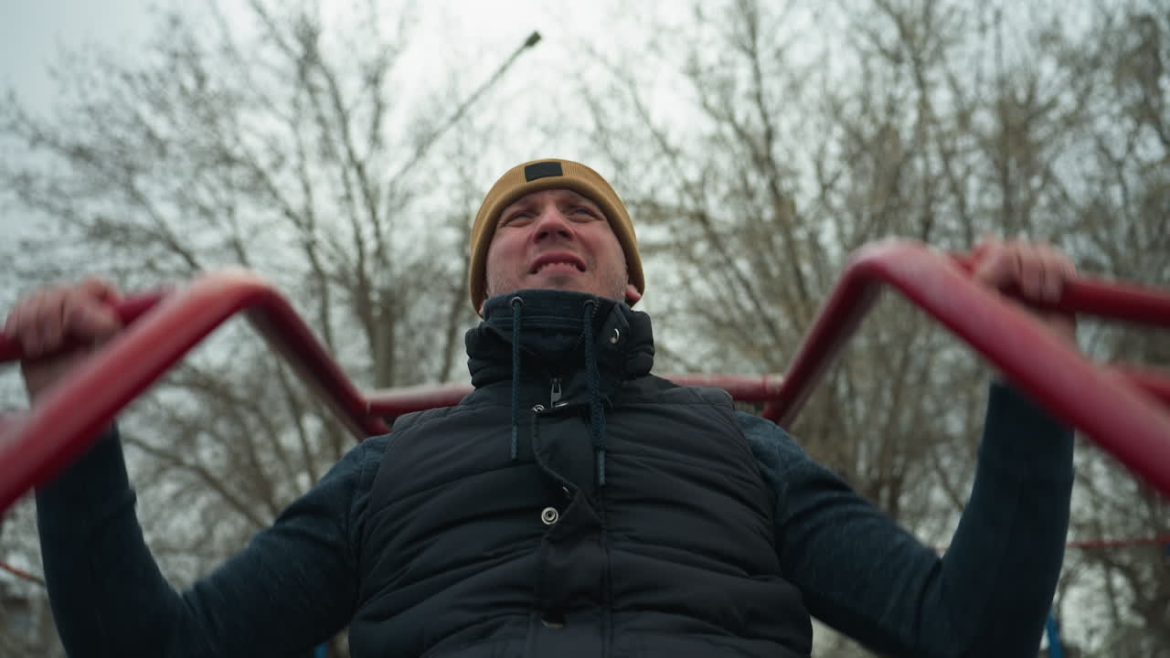 A coach wearing a brown beanie and black jacket is working out on outdoor equipment, gripping red iron bars while doing pull-ups, looking exhausted