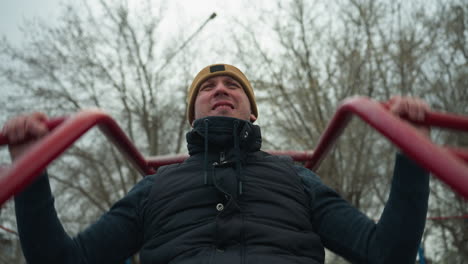 a coach wearing a brown beanie and black jacket is working out on outdoor equipment, gripping red iron bars while doing pull-ups, looking exhausted