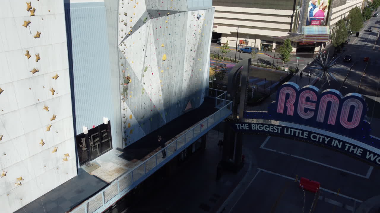 An aerial shot captures a woman rock climbing in downtown Reno, Nevada. As the camera pulls back, the view expands to reveal the city streets below, showcasing the iconic Reno sign.