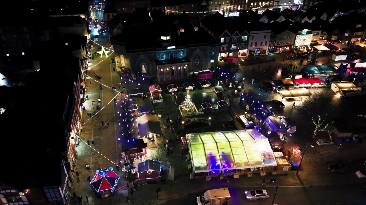 Flygfoto med drönare på natten över Salisburys julmarknad som visar Guildhall, upplysta stånd, en dekorerad gran och ett livligt marknadstorg.