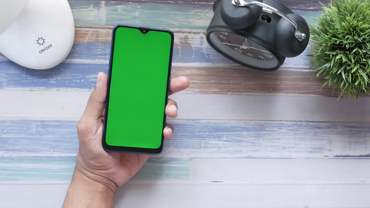 persona sosteniendo un teléfono inteligente con una pantalla verde en un escritorio con una lámpara y un despertador