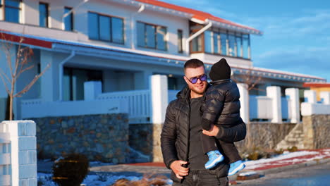 happy smiling man holding a little baby boy. father and son wearing sunglasses and warm clothes stand near the house outdoors.