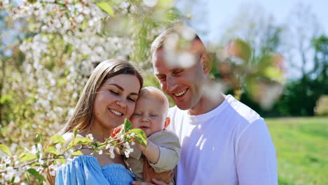 loving mom and dad kiss their beloved adorable baby on the head. little child tries to grab a tree branch in bloom. happy family outdoors.