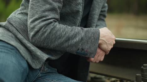 una vista de cerca de un hombre con un blazer gris y pantalones vaqueros azules, sentado y juntando las manos en una vía ferroviaria