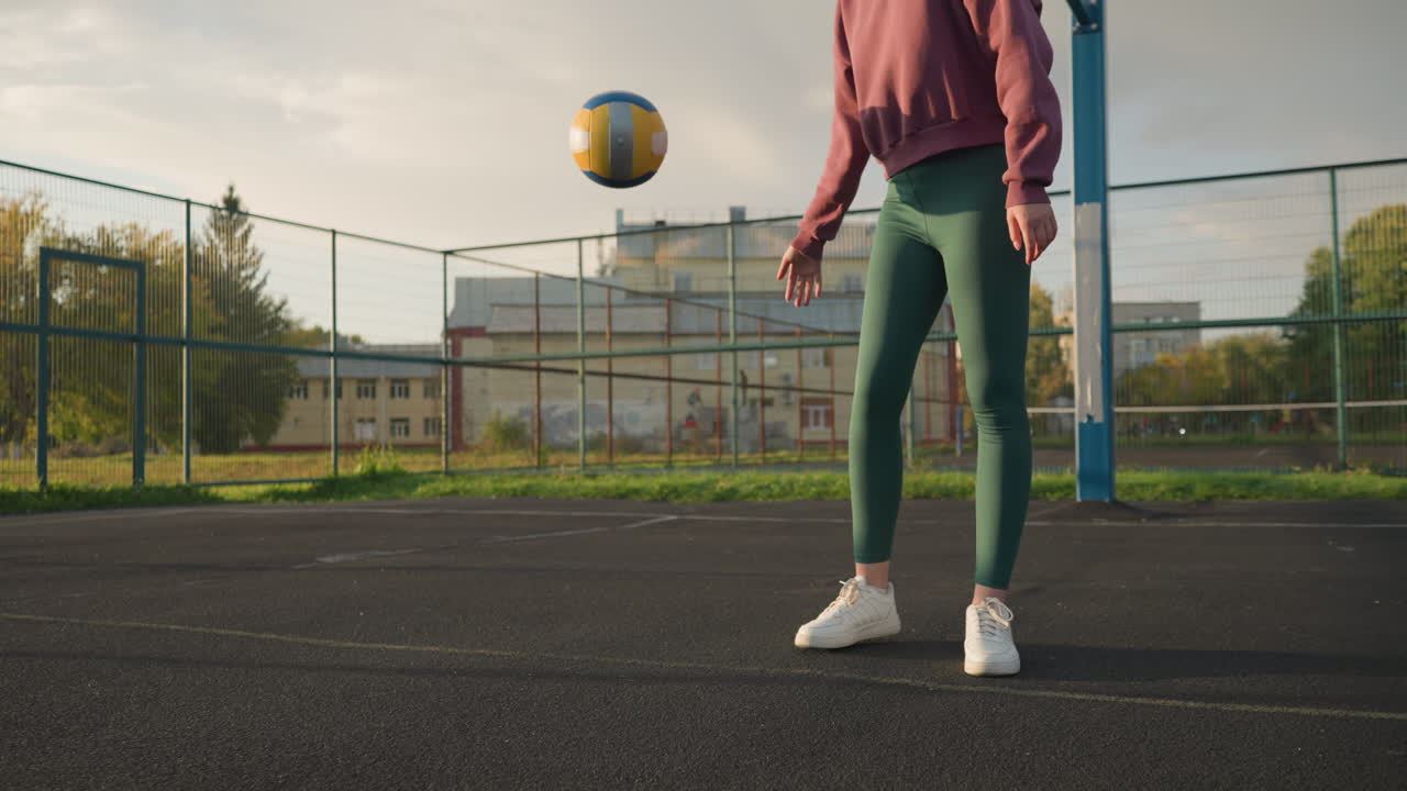 Young athletic woman in sportswear bouncing volleyball on outdoor court with basketball hoop and fenced sports area, background features a building and lush greenery