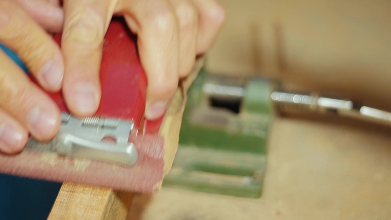 Close-up of a craftsman using a red sanding block to smooth a wooden plank by hand in a workshop. Authentic DIY woodworking footage for craft, carpentry and maker projects