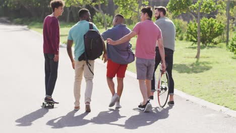 felices amigos varones diversos caminando en bicicleta y hablando en un día soleado