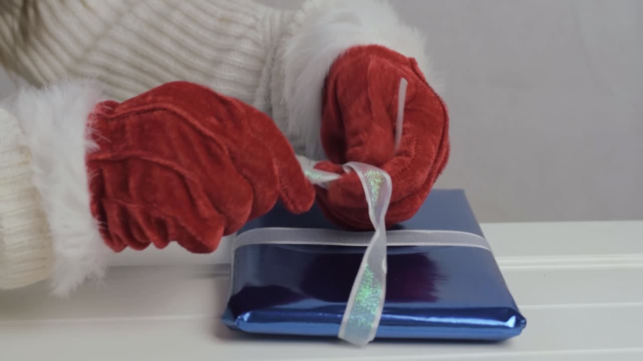 Hand wearing Santa gloves wrapping Christmas gift with ribbon and blue paper medium shot