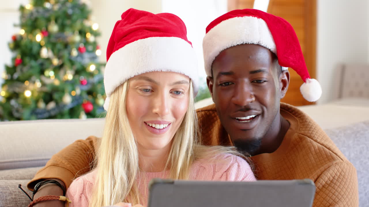 diverse couple wearing Santa hats enjoying tablet together by Christmas tree