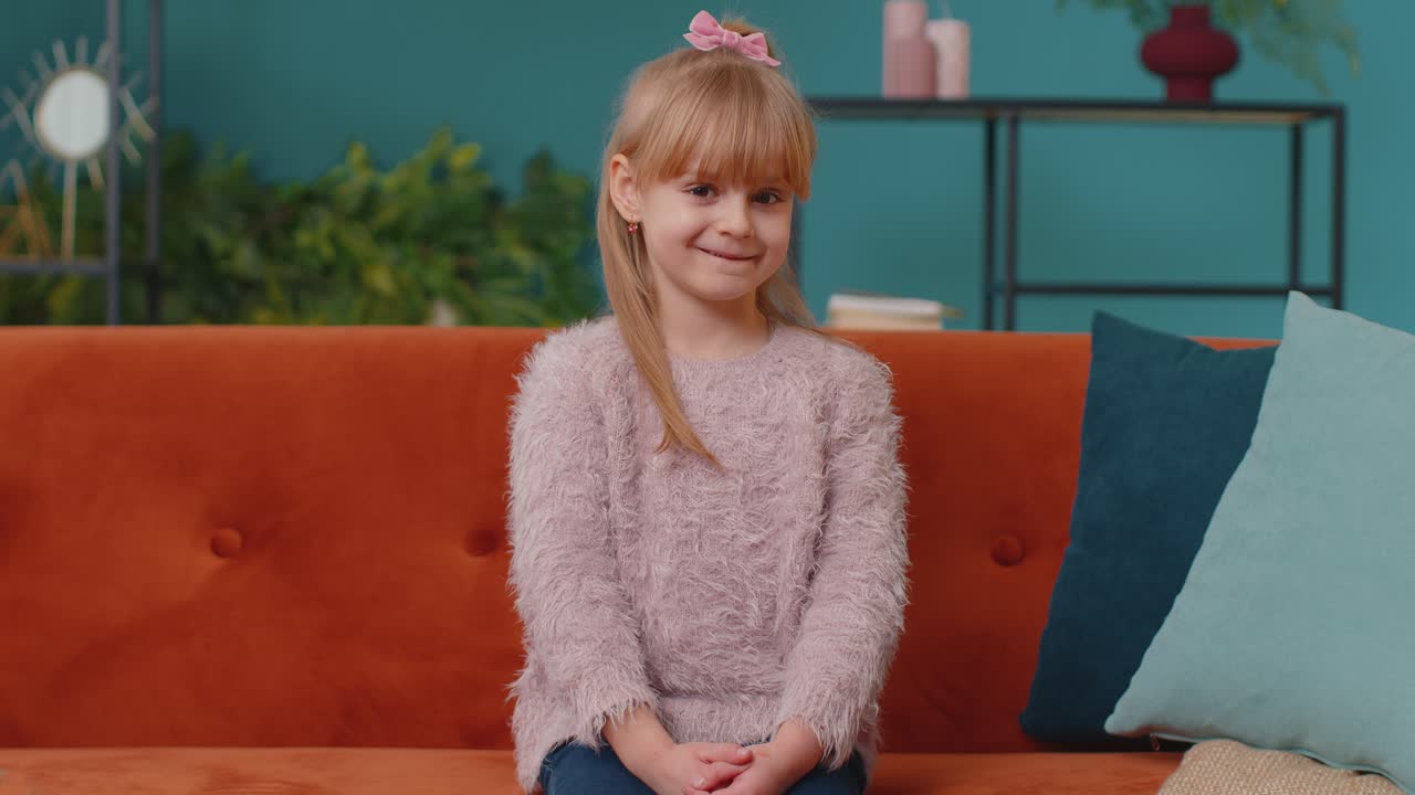 Portrait of little adorable child girl sitting alone on sofa at home looking at camera and smiling