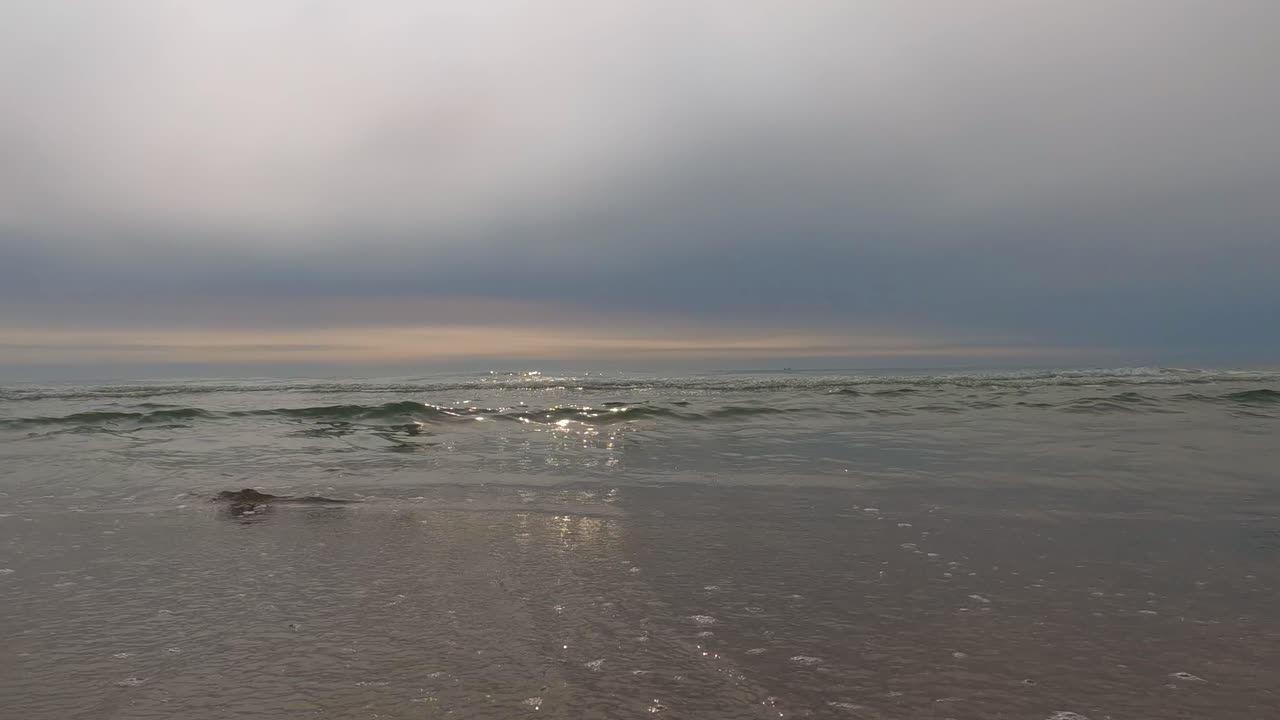 Low-angle view of gentle waves rolling onto Smiltynė beach under a moody sky, with soft light reflecting on the Baltic Sea