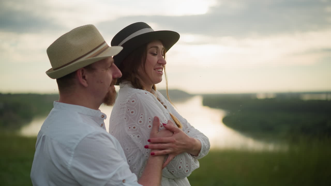 A romantic moment captured between newlyweds dressed in white, both wearing hats, as they stand close to a serene lakeside at sunset. The man lovingly holds the woman from behind