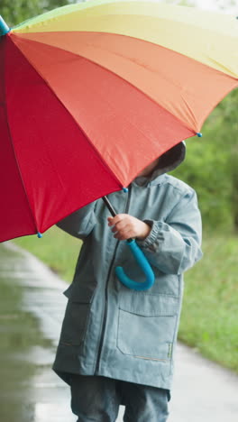 child twirls umbrella in hands in park. boy plays with multicolored parasol spending time in garden in rainy day. mesmerizing whirl among rain-soaked air