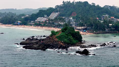 tropical sri lanka slightly elevated view of a rocky islet covered in green foliage, near a sandy beach and bustling coastal town under an overcast, humid sky, captured with a standard lens