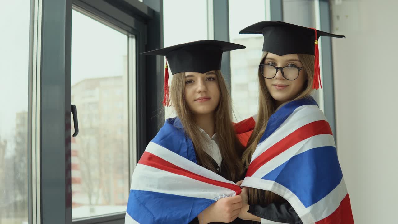 Two young happy graduate women stand shoulder to shoulder with a British flag on their shoulders
