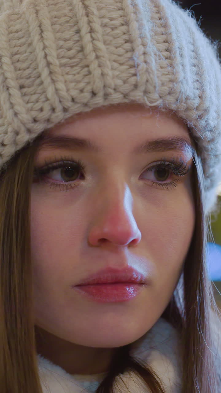 Close-up of young woman wearing a knitted beanie and winter coat, sitting alone with eyes closed in deep reflection, blurred festive city lights and people moving in the background