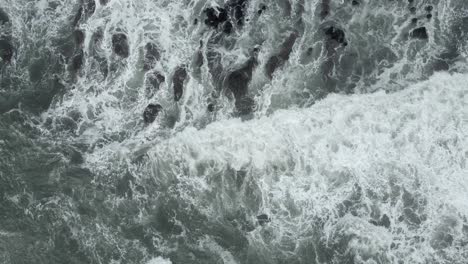 Grey-green-ocean-water-breaks-in-white-foam-rocks-near-shore,-aerial