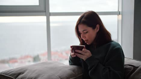 A-brunette-girl-in-a-green-jacket-sits-on-the-sofa-holds-a-pillow-on-her-legs-and-drinks-hot-tea-from-a-brown-mug-in-a-modern-apartment-overlooking-the-sea