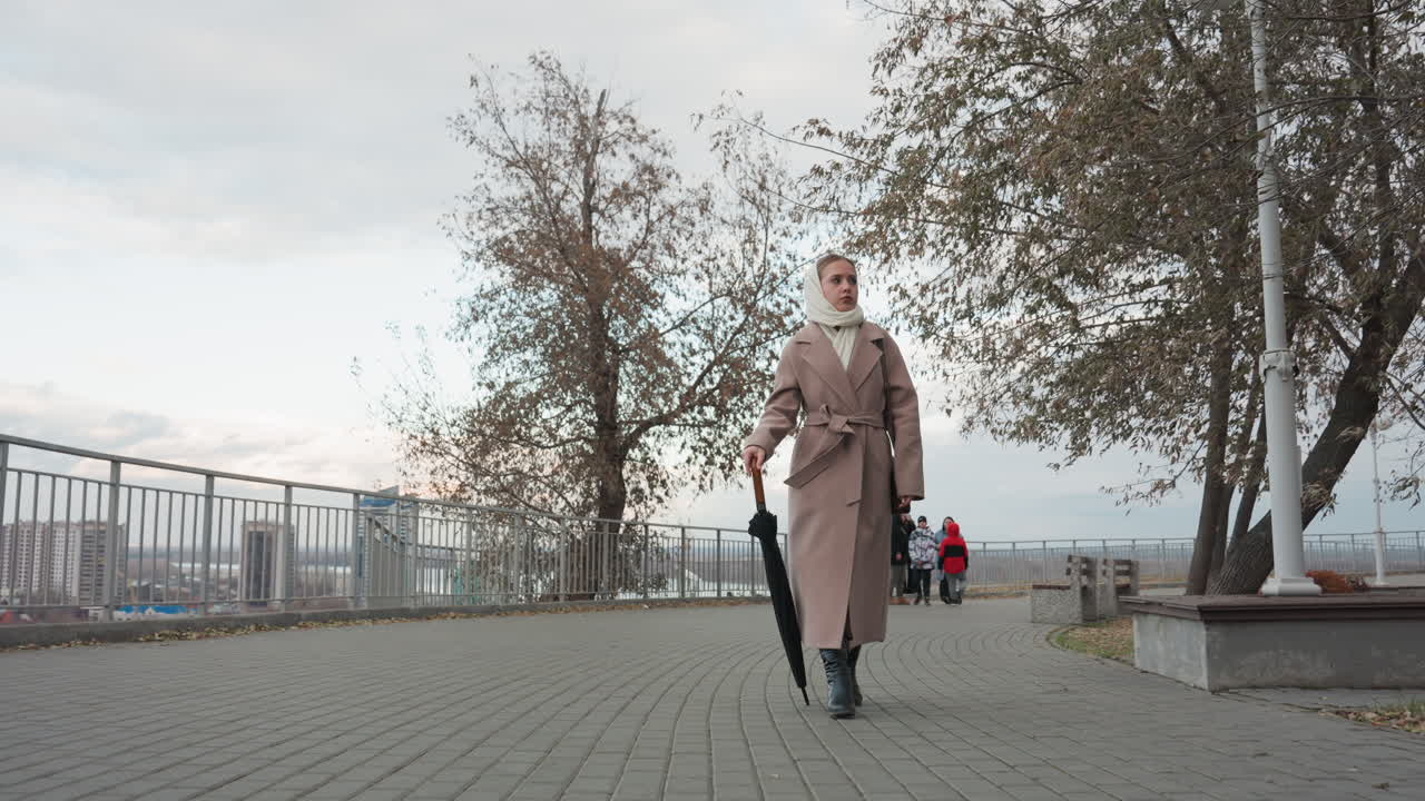 White woman walking waterfront promenade, beige trench coat and closed umbrella, overcast sky, autumn leaves on pavement, distant harbor and skyline, lamppost and bench, slow contemplative pace, muted