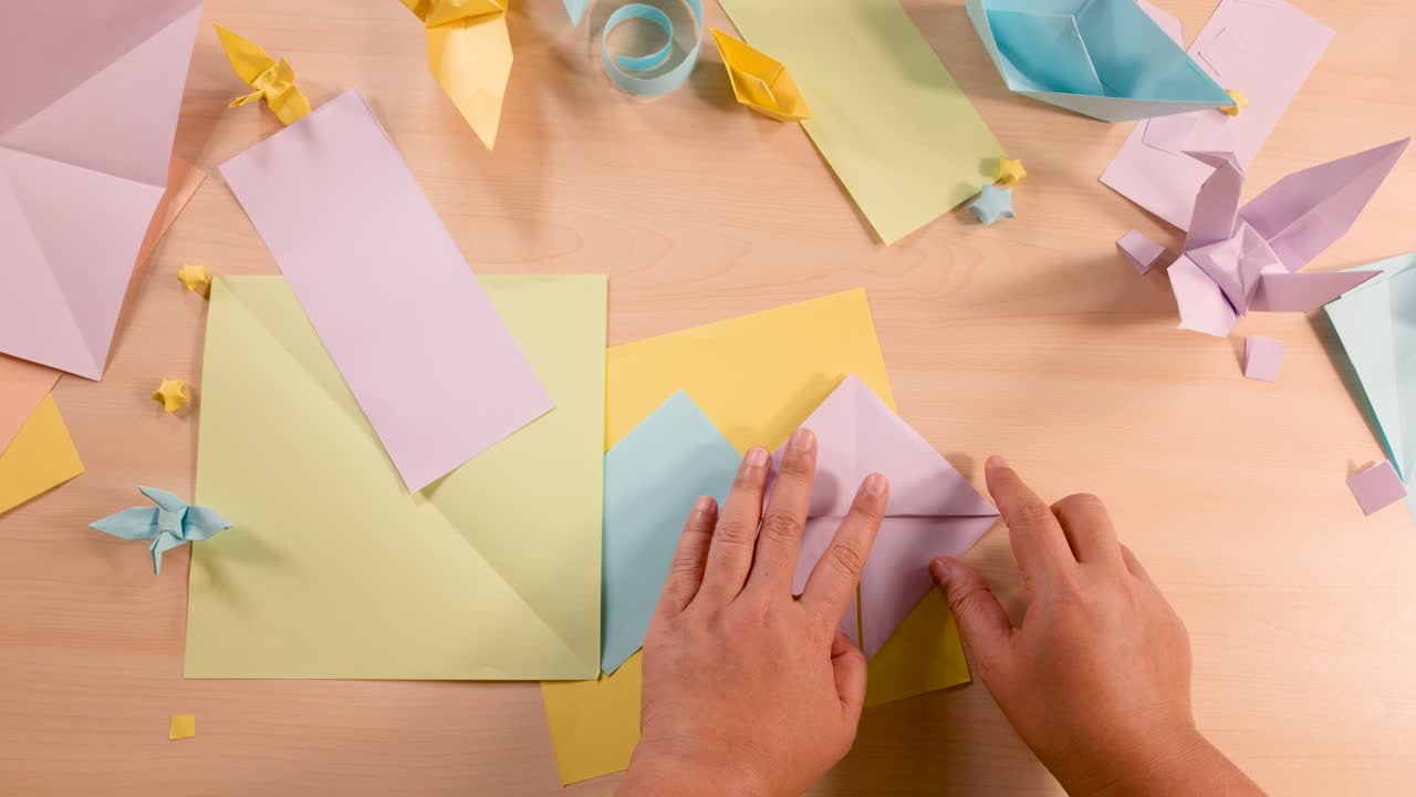 Person folds pastel origami paper, overhead view, even lighting, colorful craft environment, steady camera