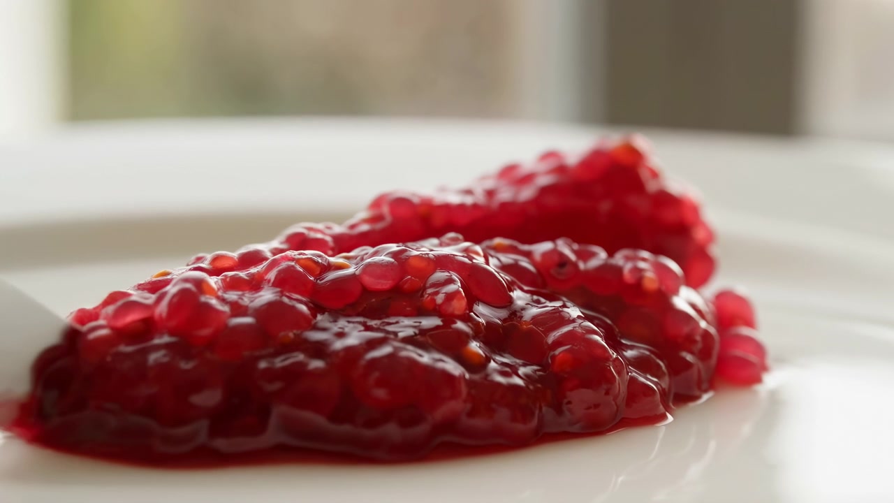 Coltello appoggiato contro polpa di melograno rossa lucida su piatto bianco, con grappoli di semi, lucentezza umida, succo sciropposo, sfondo morbido e sfocato, angolazione bassa, inquadratura ravvicinata, luce diffusa brillante, fresco e di alta qualità.