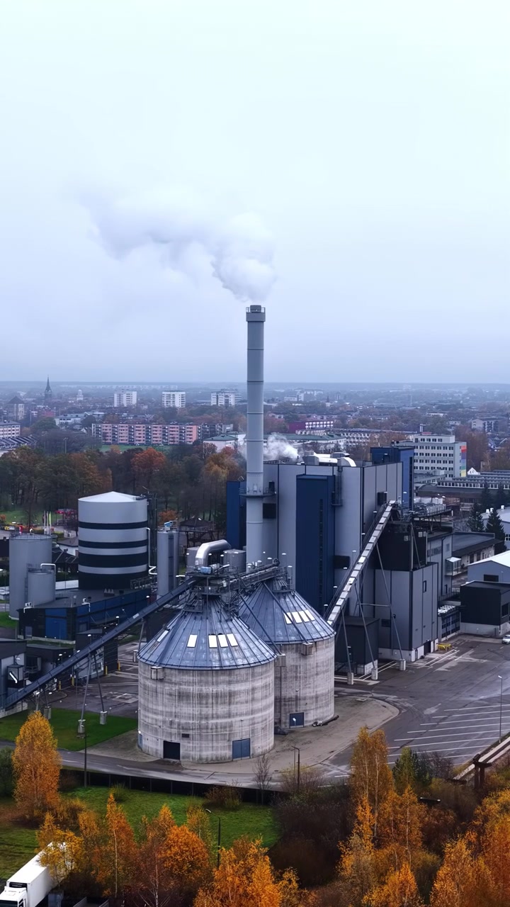 An aerial view of a modern industrial facility and heating plant with white smoke rising from a tall chimney, surrounded by autumn foliage and city buildings.