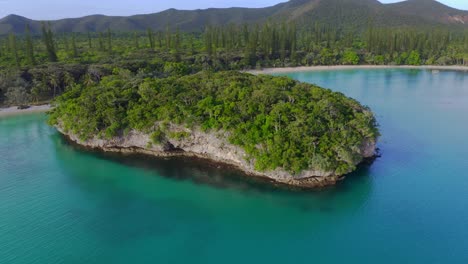 Drone-shot-flying-backward-from-a-natural-rock,-island-formation-to-reveal-the-landscape-with-mountains,-forest,-beach-and-a-blue-lagoon-during-a-beautiful-day