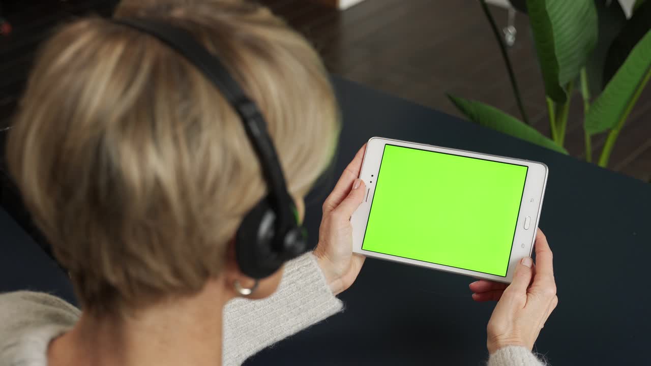una mujer de mediana edad con auriculares está viendo contenido usando una tableta en una pantalla verde, llamando a sus parientes