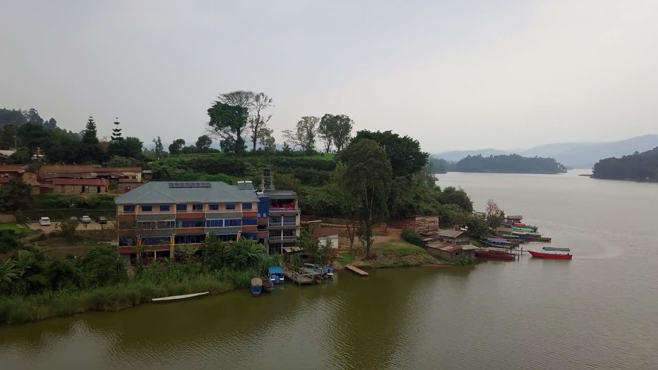 Top-down aerial of a tranquil fishing market on Lake Bunyonyi, Uganda, showing moored boats, small docks, waterfront buildings with solar roofs and surrounding trees against calm reflective water