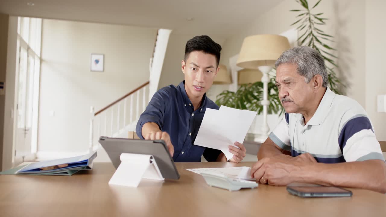 Diverse financial advisor and senior man discussing paperwork and using tablet at home, slow motion