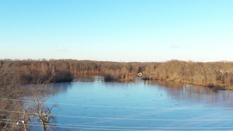 an aerial view of a quiet lake with dry leafless trees surrounding it on a sunny evening