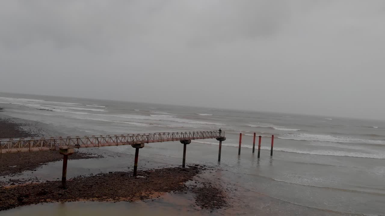 Person walking on a pier at Allana Gadoor Village Beach under an overcast sky