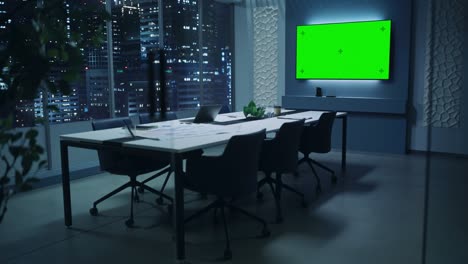 modern empty meeting room with big conference table with various documents and laptop on it. wall tv showing green chroma key screen mock up display template. late evening or night city outside.