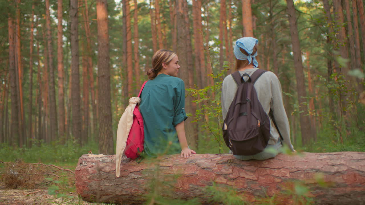 dos excursionistas comparten un momento alegre en un árbol caído en un bosque pacífico, una con bandana azul y mochila negra sonríe cálidamente, mientras que la compañera con camisa verde y bolsa roja limpia su mano después de sentarse