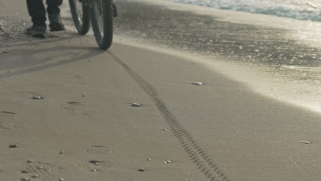 a man walks alongside beach with his bicycle, leaving footprints and tire tracks in the wet sand