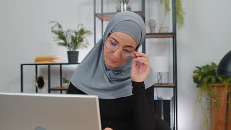 Muslim-business-woman-in-hijab-looking-at-camera-after-finishing-laptop-computer-work-at-home-office