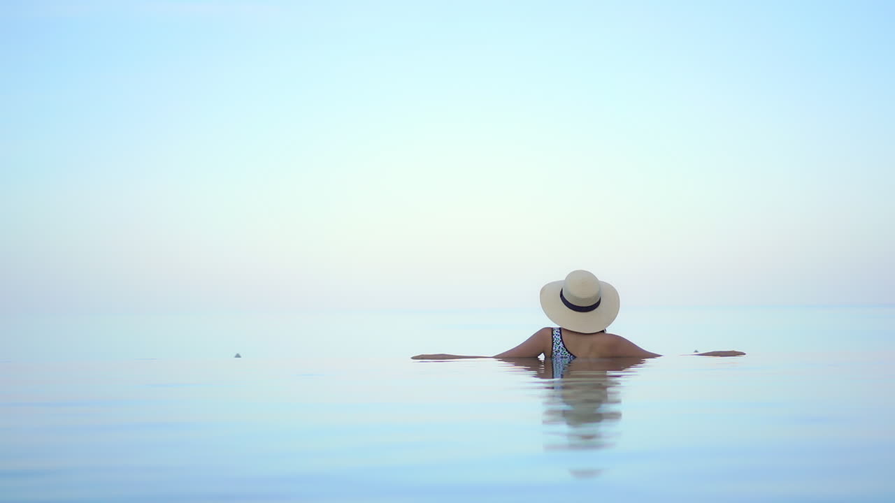 끝없는 바다 전망을 내다보는 리조트 인피니티 풀에 있는 젊은 여성을 다시 카메라로